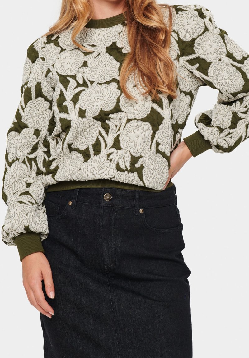 Olivgrüner gestickter Pullover mit weißen Blumenmuster, strukturiertem Stoff, gerippten Bündchen und Saum. Kombiniert mit einem dunklen Jeansrock.