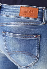 Primer plano de un bolsillo trasero de unos jeans de mezclilla azul con una etiqueta de cuero marrón claro que dice "Hilfiger Denim, Un Original Americano."