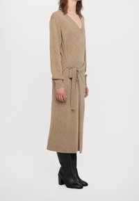 Robe tricotée beige avec un profond décolleté en V, des manches longues et un détail de taille nouée. Elle présente une texture côtelée et une longueur midi. Portée avec des bottes à talons noires.