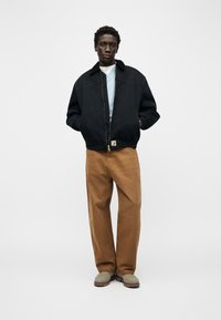 Άνδρας στέκεται με τα χέρια στις τσέπες, φοράει μαύρο μπουφάν, γαλάζιο πουκάμισο, καφέ παντελόνι και μπεζ μπότες, πάνω σε λευκό φόντο.