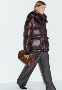 Persona bionda che cammina, indossando una giacca a vento lucida scura, pantaloni grigi a gamba larga, scarpe nere, e tiene in mano una borsa a mano in pelle marrone.