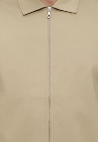 Κοντινή λήψη ενός μπεζ σακακιού με γιακά και ασημί φερμουάρ ολόκληρου μήκους στο κέντρο.