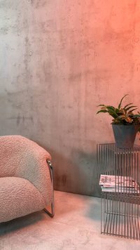 Καμπυλωτή μπεζ πολυθρόνα δίπλα σε μεταλλικό τραπεζάκι με σύρμα που κρατά περιοδικά και μια γλάστρα απέναντι από μια υφή σκυρόδεματος.