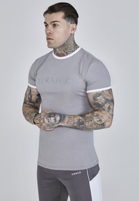 Grå tætsiddende t-shirt med en hvid ribkant, der har et broderet "SIKSILK" logo på brystet. Matcher med grå og hvide træningsbukser.