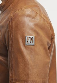 Brun læderjakke med en struktureret finish, der viser et metal logo-badge på skulderen og detaljeret syning langs sømmene.