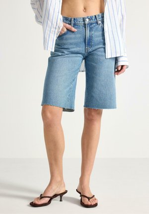 Kvinde iført lyseblå højtaljede denim Bermudashorts, stribet hvid skjorte og brune hælsandaler, stående mod ensfarvet baggrund.