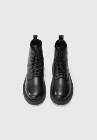 Mustat nahkaiset nilkkasaappaat, joissa on kohokuvioidut sivut, paksut pohjat, nauhat ja vetolenkit takana, kuvattu ylhäältä valkoista taustaa vasten.