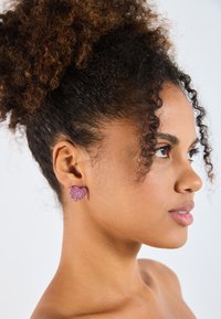 Pendientes rosa en forma de corazón con un acabado brillante, llevados en la oreja. Superficie texturizada, de pequeño tamaño y con un respaldo suave.