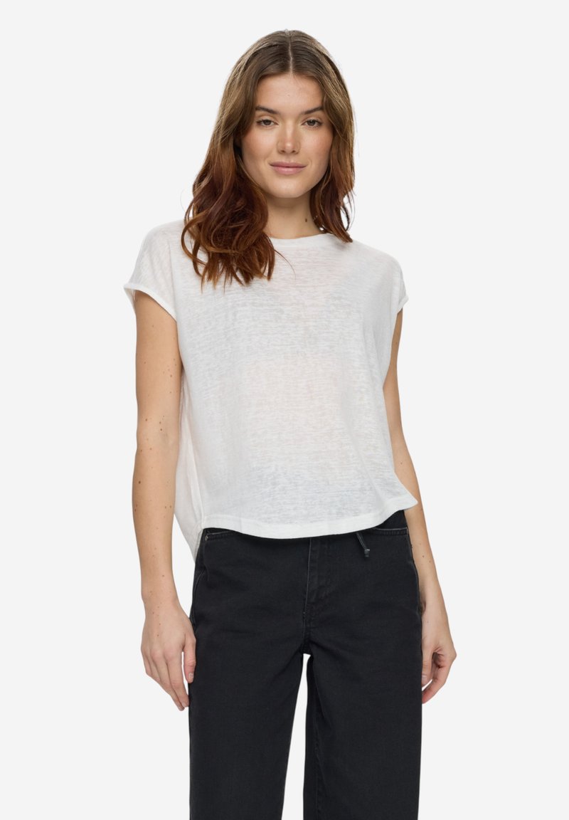Femme aux cheveux bruns ondulés, portant un haut blanc ample à manches courtes et un pantalon noir taille haute, debout devant un fond uni.