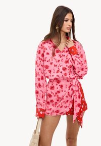 Femme portant une blouse rose à fleurs avec des poignets rouges à motifs floraux et une jupe portefeuille assortie, tenant un sac à main tissé beige.