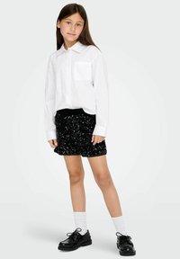 Baltas ilgomis rankovėmis marškinėliai su krūtinės kišenėle, derinami su juoda blizgių vakarėlių mini sijonu ir juodais blizgiais batais. Baltos kojinės užbaigia aprangą.