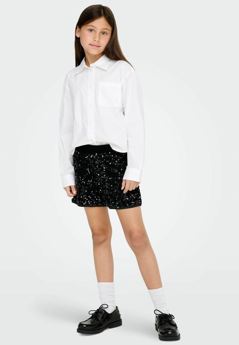 Baltas ilgomis rankovėmis marškinėliai su krūtinės kišenėle, derinami su juoda blizgių vakarėlių mini sijonu ir juodais blizgiais batais. Baltos kojinės užbaigia aprangą.