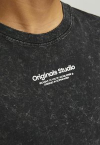 Juodas medvilninis marškinėliai su banguotu apvaliu kaklu, su nusidėvėjusia tekstūra ir baltais užrašais "Originals Studio" priekyje.