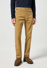 Pantaloni in corduroy beige con vestibilità dritta, dotati di tasche anteriori e un piccolo logo sul punto vita. Abbinati a scarpe marroni.