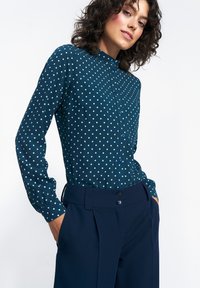 Granatowa koszula z długim rękawem w jasnoniebieskie kropki, zapinana na guziki, z wysokim kołnierzem i miękkiego materiału. Połączona z dopasowanymi ciemnymi spodniami.