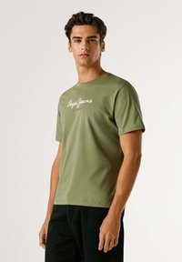 Alyvuogių žalios spalvos apvalios apykaklės marškiniai iš medvilnės audinio, su balta skirta užrašyta "Pepe Jeans London". Trumpomis rankovėmis, laisvu siluetu. Juodos kelnės.