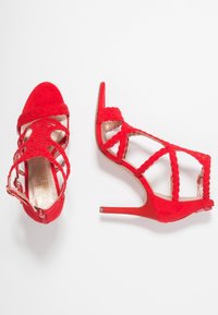 Červené sandály na podpatku s pletenými pásky, otevřenou špičkou a strukturovaným surovým vzhledem. Mají štíhlý stiletto podpatek a zip na kotníku.