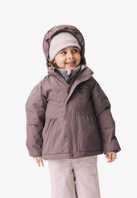 Lilla isolert jakke med hette, glidelås og sidelommer, båret over et grått fleece lag og lyse bukser. Accentert med en beige strikket lue.