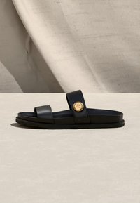 Μαύρο σανδάλι με δύο λουράκια, το ένα με στρογγυλό χρυσό έμβλημα, πάνω σε ουδέτερο μπεζ φόντο.