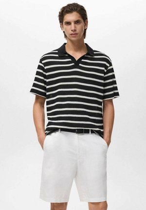 Young man stands confidently, wearing a black and white striped polo and white shorts, with hands in pockets, against a plain background.