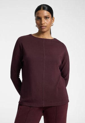 Femme aux cheveux foncés attachés en arrière porte un haut à manches longues et un pantalon assortis en bordeaux profond, debout, les mains derrière le dos.
