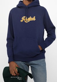 Man som bär en marinblå hoodie med gul "Ralph" text, ljusblå jeans, håller en mörkgrön keps med broderad Polo Bear.