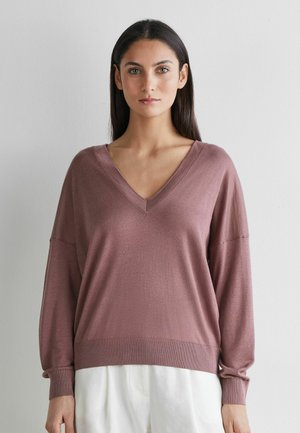 Kvinde med langt mørkt hår iført en mauve V-hals sweater og hvide bukser, stående mod en ensfarvet lys grå baggrund.