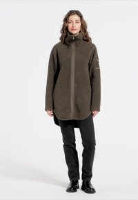 Olivengrüne Fleece-Jacke mit Reißverschluss, hohem Kragen und langen Ärmeln. Verfügt über einen seitlichen Schlitz und eine strukturierte Oberfläche. Getragen mit schwarzen Hosen und Stiefeln.