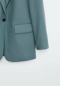 Blazer sarcelle à un bouton avec poche à rabat sur un fond clair uni, montrant la partie inférieure droite devant et la manche.