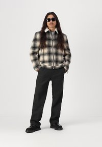 Juodos ir baltos languotos zip-up striukės su kontrastinga balta apykakle, derinamos su tamsiomis kelnėmis ir juodais batais. Modelis nešioja saulės akinius.