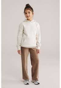 Ragazza giovane con capelli scuri raccolti in uno chignon, indossa una felpa crema con cappuccio, pantaloni di velluto a coste marroni e scarpe da ginnastica bianche, in piedi davanti a uno sfondo beige.