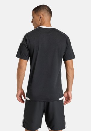 Mężczyzna ubrany w czarną sportową koszulkę i szorty z białymi akcentami, stojący tyłem na jednolitym jasnym tle.