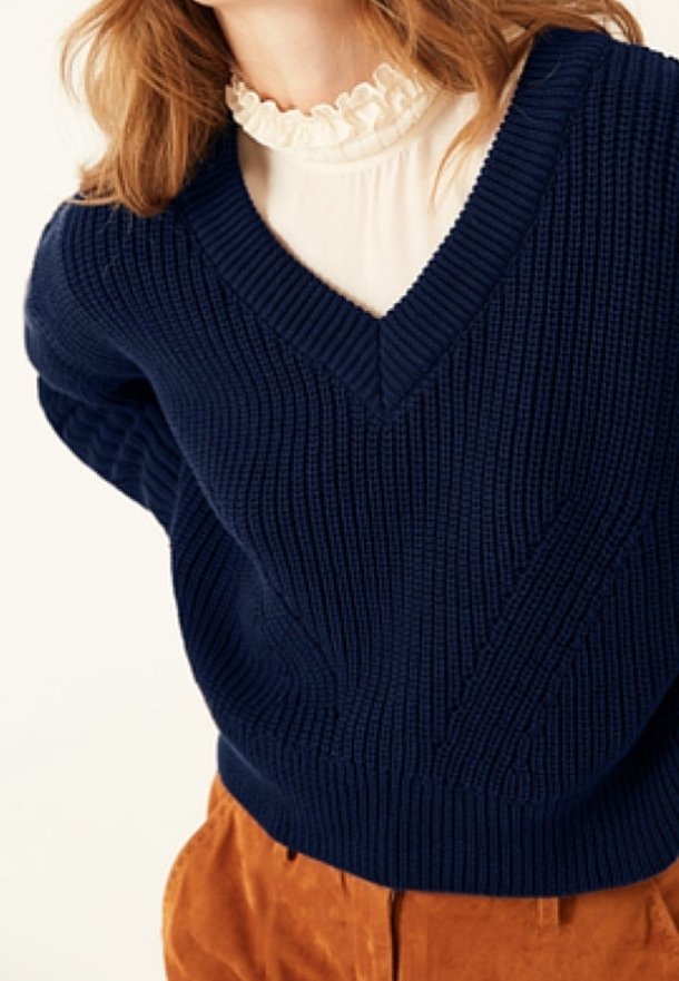 Kobieta w granatowym swetrze z dekoltem w serek, białej bluzce z falbanami przy wysokiej szyi oraz w rdzawych spodniach, widoczna od pasa w górę.