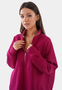Sudadera fucsia con cremallera y puños acanalados, con un cuello alto, que presenta una textura suave y un ajuste relajado. Detalle de cremallera metálica.