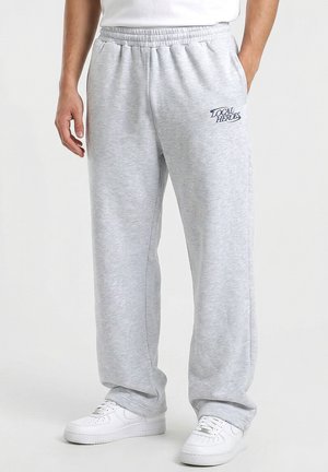 Mężczyzna w jasnoszarych spodniach dresowych z logo "Local Heroes" na lewej uda i białych tenisówkach, stojący z rękami swobodnie opuszczonymi.