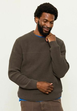 Brauner gerippter Strickpullover mit Rundhalsausschnitt, strukturierter Stoff und langen Ärmeln. Über einem blauen Hemd getragen. Taillierte Passform.