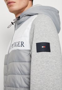 Lichtgrijze hoodie met een puffy witte en grijze voorkant, voorzien van een logo op de borst en een marineblauw merkenlabel op de linkermouw.