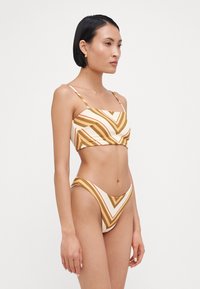 Dvoslojni bikini komplet s patterned zgornjim delom z rjavimi in kremnimi zigzag črtami ter ustreznimi visokoreznimi hlačkami. Material se zdi teksturiran.