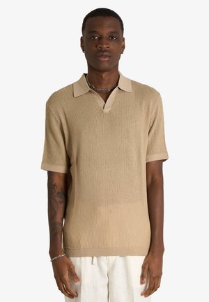 Uomo che indossa una polo beige a maniche corte in maglia e pantaloni bianchi, in piedi con le braccia rilassate, espressione neutra, sfondo bianco uniforme.