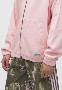 Άτομο φοράει ένα ανοιχτό ροζ μπουφάν Adidas Originals με φερμουάρ και πράσινο χακί παντελόνι με ροζ ρίγες στα πλαϊνά.
