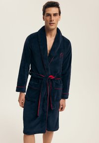 Granatowy szlafrok z polarowego materiału z czerwonymi akcentami, z kołnierzem szalowym, paskiem i dwiema przednimi kieszeniami. Gładka textura i długość do połowy łydki.