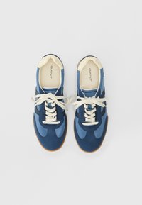 Pár modro-námořnických šněrovacích tenisek s bílými podrážkami a krémovými akcenty, z pohledu shora na bílém pozadí.