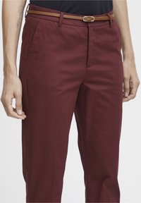 Pantalon bordeaux en tissu lisse, présentant une coupe droite, des passants de ceinture et une ceinture en cuir marron avec une boucle métallique.