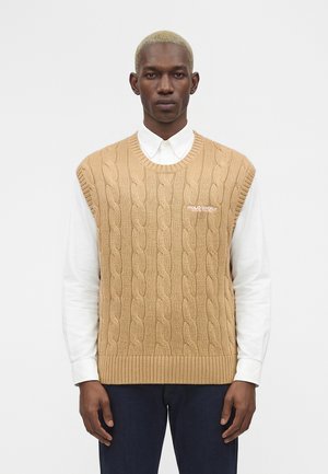 Muž s krátkymi blond vlasmi nosiaci béžový bezrukávový sveter s vrkočovým vzorom cez bielu zapínaciu košeľu a tmavé nohavice na jednofarebnom pozadí.