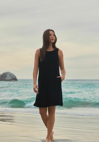 Robe noire sans manches en tissu léger, dotée de poches et d'une coupe ample, posée contre un fond de plage avec des vagues douces.