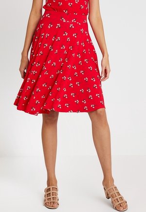 Vrouw draagt een rode bloemenprint A-lijn rok en beige sandaal met blokhak en bandjes, staand tegen een effen achtergrond.