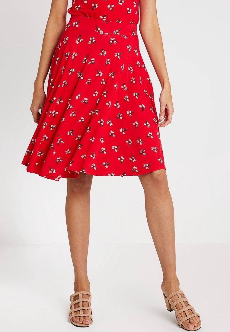 Femme portant une jupe rouge à fleurs en ligne A et des sandales beige à talons blocs avec brides, se tenant devant un fond uni.