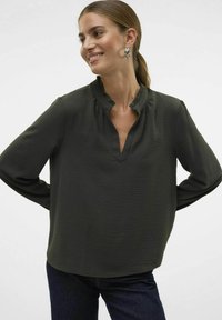 Donkergroene blouse met een ruches bij de halslijn, lange mouwen en een geweven stof. Heeft een V-vormige uitsnijding bij de kraag en subtiele plooidetails.