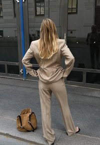 Beżowy garnitur o dopasowanym kroju z wyraźnymi ramionami, pionowymi szwami i szerokimi spodniami. W pobliżu na ziemi leży brązowa torba.