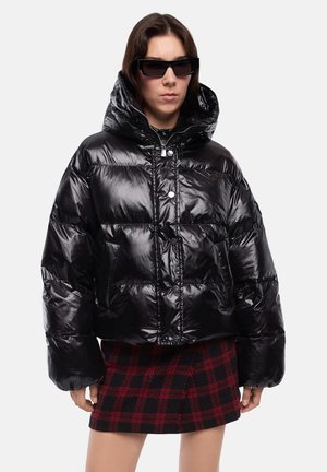 Donna che indossa una giacca a piumino nera lucida oversize con cappuccio, occhiali da sole neri e una gonna a quadri rossa e nera su sfondo neutro.
